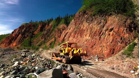 Large-powerful-yellow-mining-bulldozer-operating-and-driving-on-brown-dirt-unpaved-rocky-remote-rural-road-by-red-bedrock-hills-and-green-Yukon-landscape,-Canada,-aerial-tracking