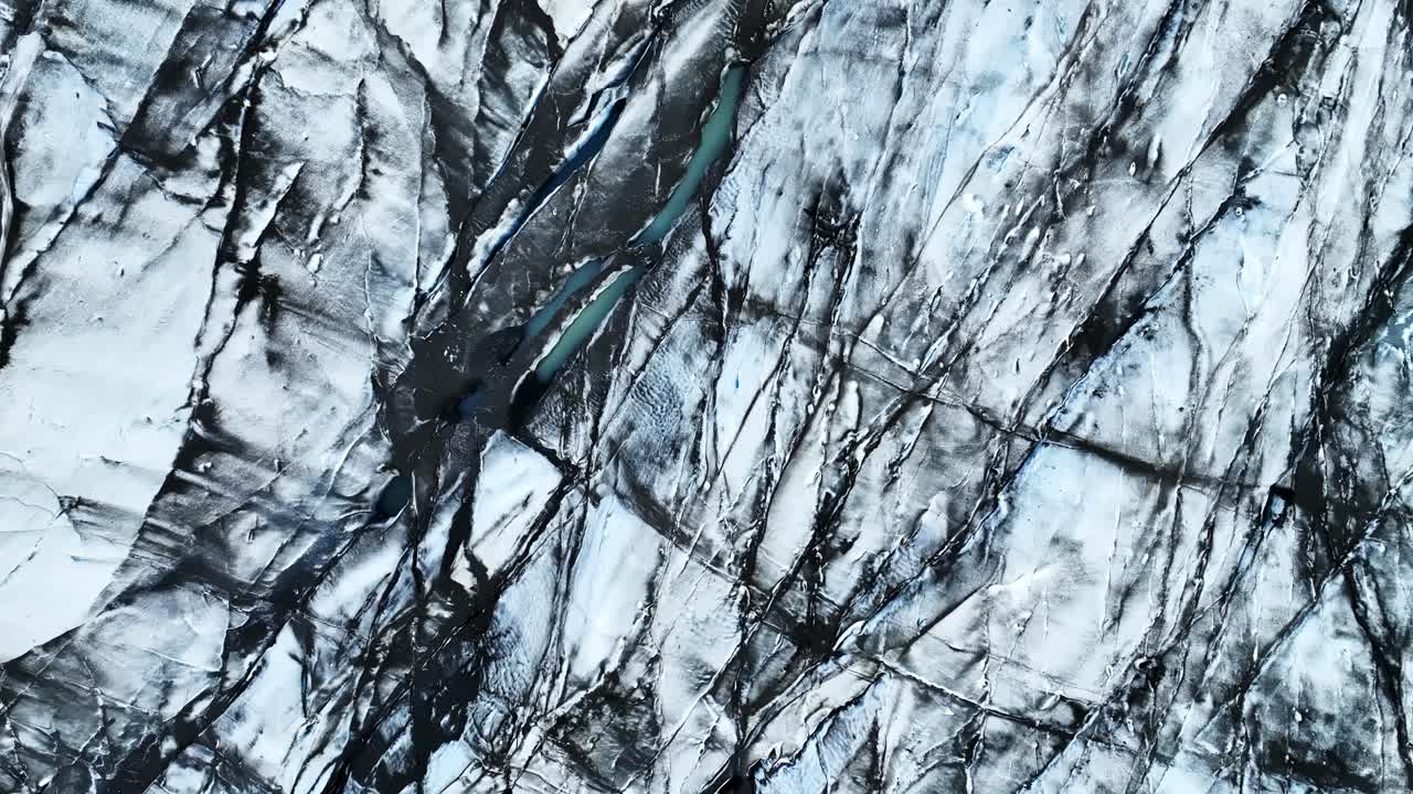 A top-down drone view over Vatnajökull reveals fractured glacier ice marked by dark volcanic lines and thin meltwater channels cutting through the frozen surface