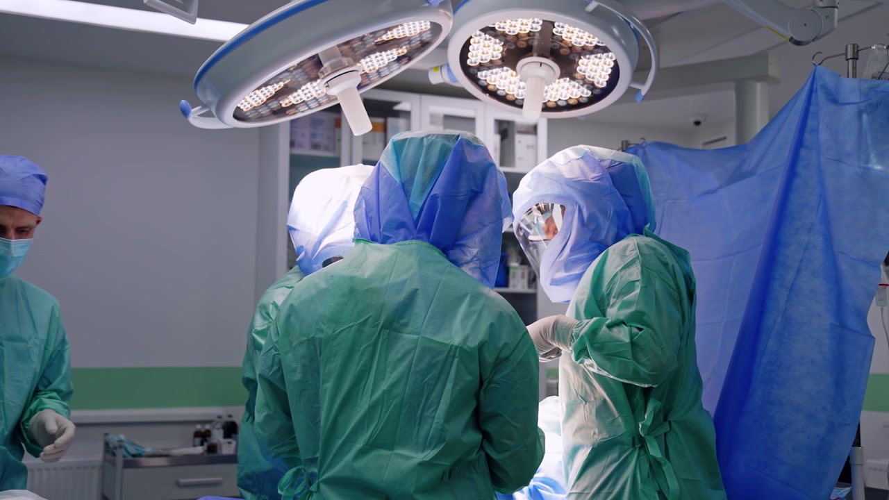 Chirurghi che collaborano con caschi protettivi lavorano in sala operatoria. I medici operano sotto luminose e moderne lampade rotonde appese al soffitto della stanza.
