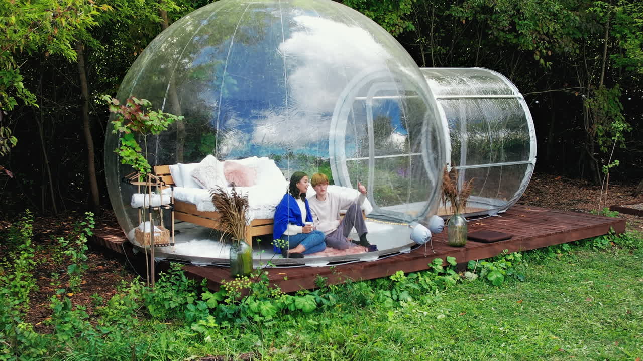 Una coppia seduta all'interno di una tenda a bolla trasparente in un sito di glamping. Seduti sotto una coperta, con delle tazze in mano mentre parlano. Foresta rigogliosa sullo sfondo.