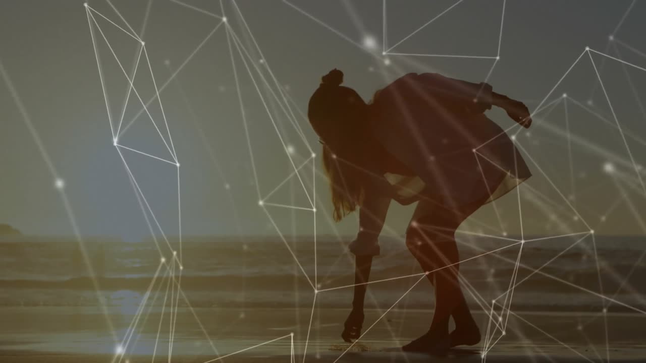 Woman pausing then picking shell while walking shoreline under digital network and dog running past