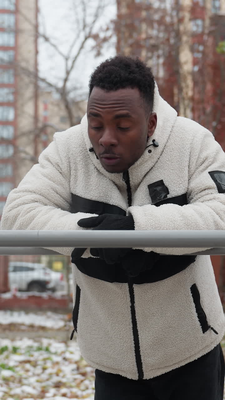 Young black man leans on iron bar, resting after workout, he looks around thoughtfully in an outdoor gym with snow-covered ground, trees, and residential buildings in the background