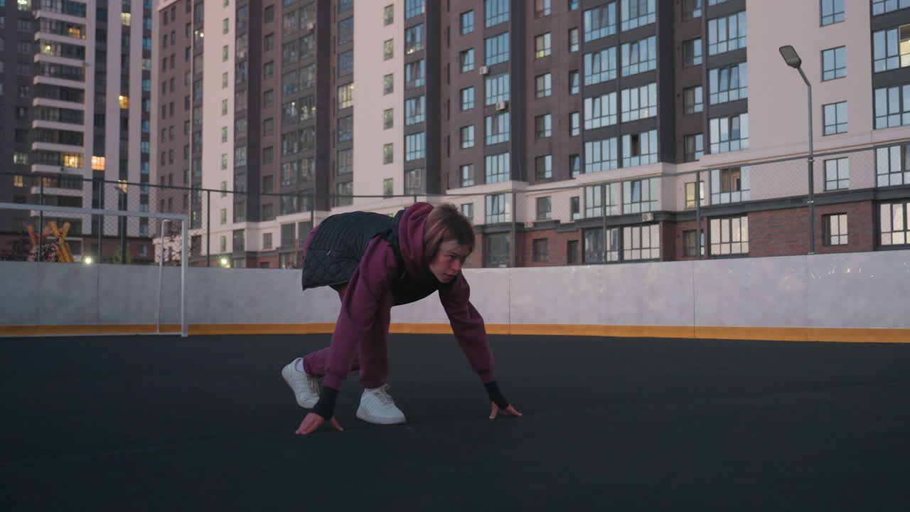 Hoodie wearing athlete crouches on white court line setting to race at dusk on black asphalt sports court near white barrier topped with chain link fence in urban high rise setting