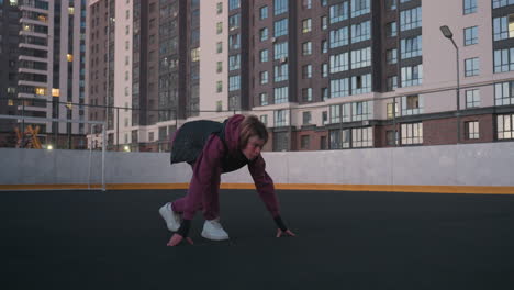 hoodie wearing athlete crouches on white court line setting to race at dusk on black asphalt sports court near white barrier topped with chain link fence in urban high rise setting