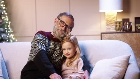 portrait of loving caring old grandfather in glasses spending christmas eve together with joyful grandchild sitting in decorated house near xmas tree embracing together