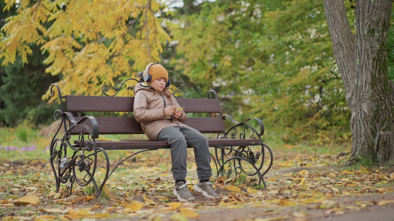 young girl seated on iron bench playing with golden leaf under yellow foliage wearing mustard beanie and headphones, surrounded by fallen leaves, absorbing music with thoughtful expression