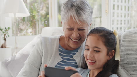 Fröhliches-Mädchen,-Das-Smartphone-Mit-Oma-Beim-Videochat-Führt,-Winkt-Der-Familie-Zu-Und-Teilt-Ihr-Urlaubswochenende-Mit-Großmutter,-Die-Auf-Dem-Handy-Chattet-Und-Sich-Zu-Hause-Mit-Enkelin-In-4K-Entspannt