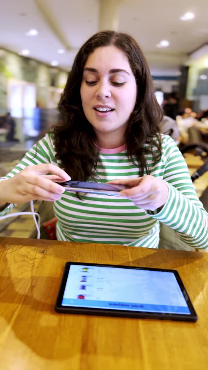 A woman is sitting at a table with a tablet and a cell phone