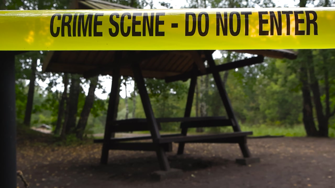 Police line yellow colored Crime Scene forensic investigation tape ribbon pulled in front of a grilling spot or outdoors forest camping ground with wooden tables and chill area during a cloudy day.