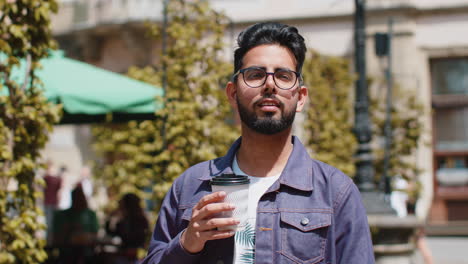 Young-indian-man-enjoying-drinking-morning-coffee-hot-drink,-relaxing,-taking-a-break-in-city-street