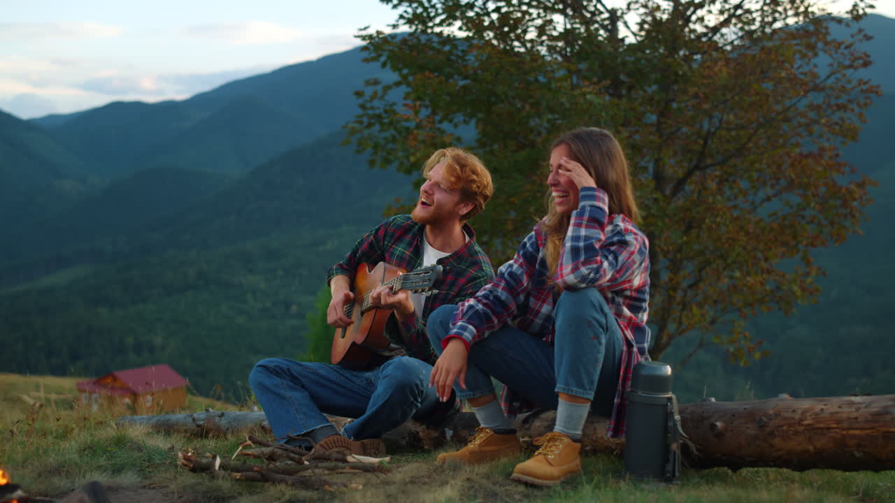 즐거운 커플이 불꽃에 가까운 자연을 즐기고 젊은 여행자들이 기타를 연주합니다.