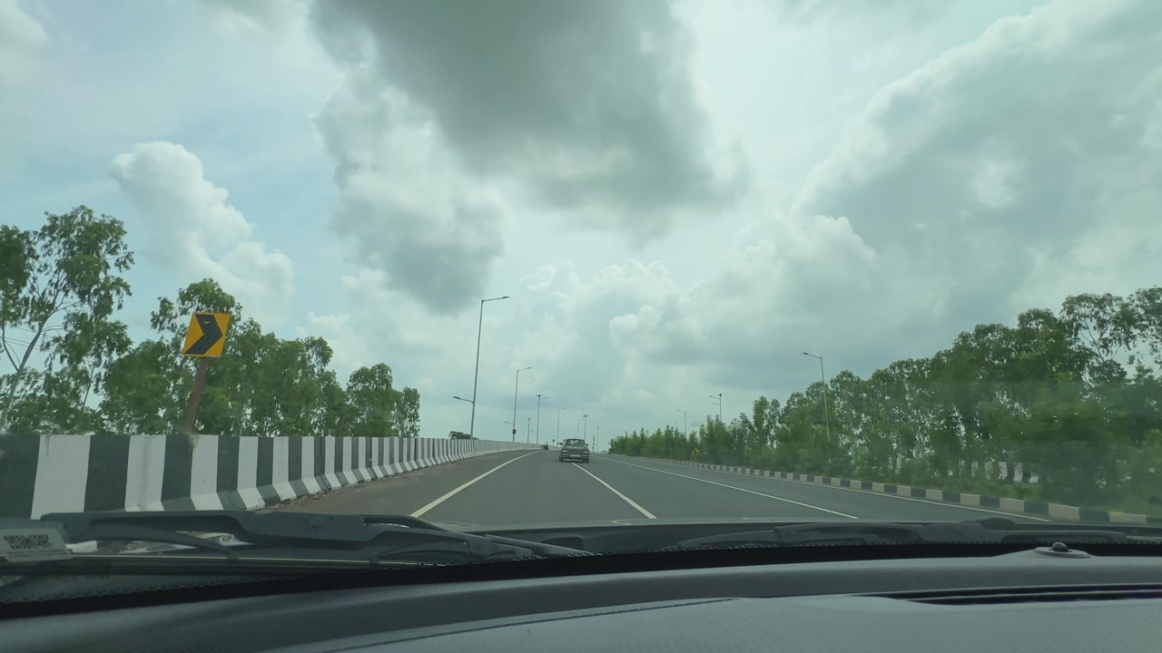 Point-of-view shot from inside a moving car on a well-constructed road in India, with trees, streetlights, and cloudy skies in view.