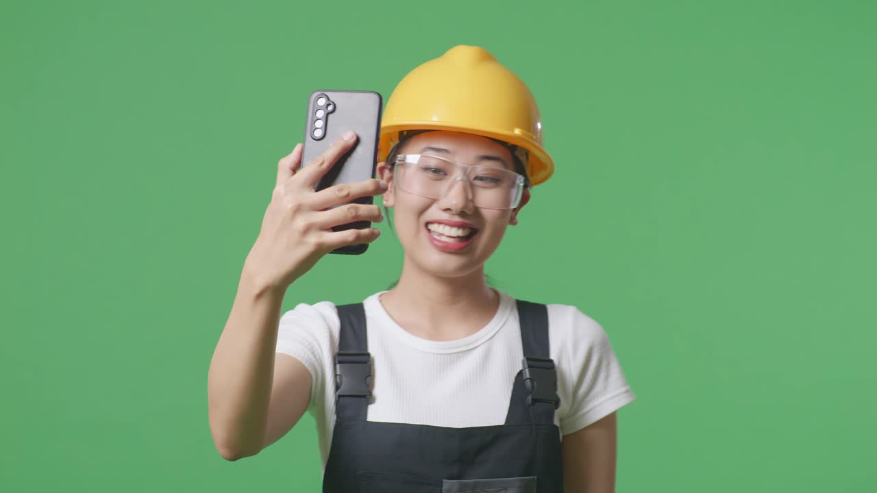 녹색 스크린 배경 스튜디오에 서서 스마트폰으로 비디오 통화를 하는 동안 손을 흔들고 보호 안경을 착용 한 아시아 여성 노동자의 근접