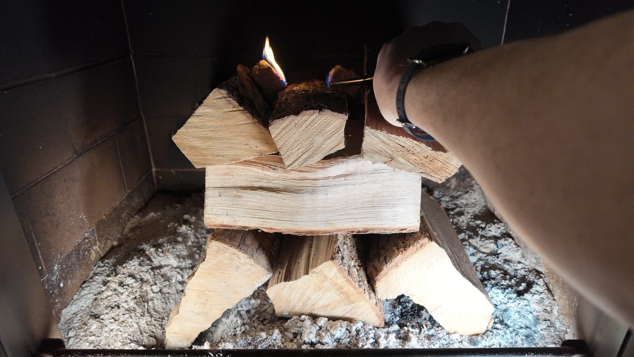 A close-up shot of a hand igniting a natural fire starter with a lighter inside a fireplace filled with stacked firewood. The flame catches on the wood shavings, symbolizing warmth, home comfort