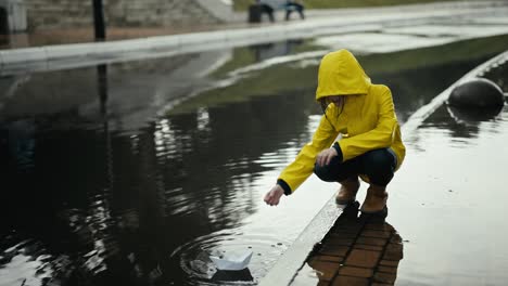 A-girl-in-a-yellow-jacket-floats-a-white-paper-boat-through-a-puddle-while-standing-on-the-curb-while-walking-in-the-drizzling-rain-on-the-street