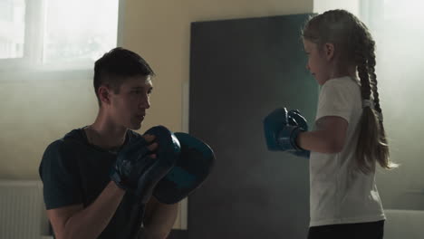 child hits pads held by trainer with hands in gym. attentive instructor trains little girl to attack in boxing school slow motion. healthy leisure