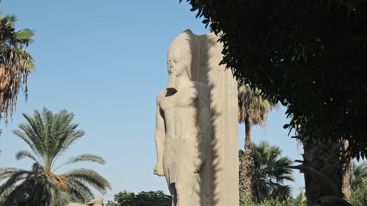 A colossal limestone statue of the pharaoh Ramesses II stands upright and prominent, wearing the distinct White Crown of Upper Egypt