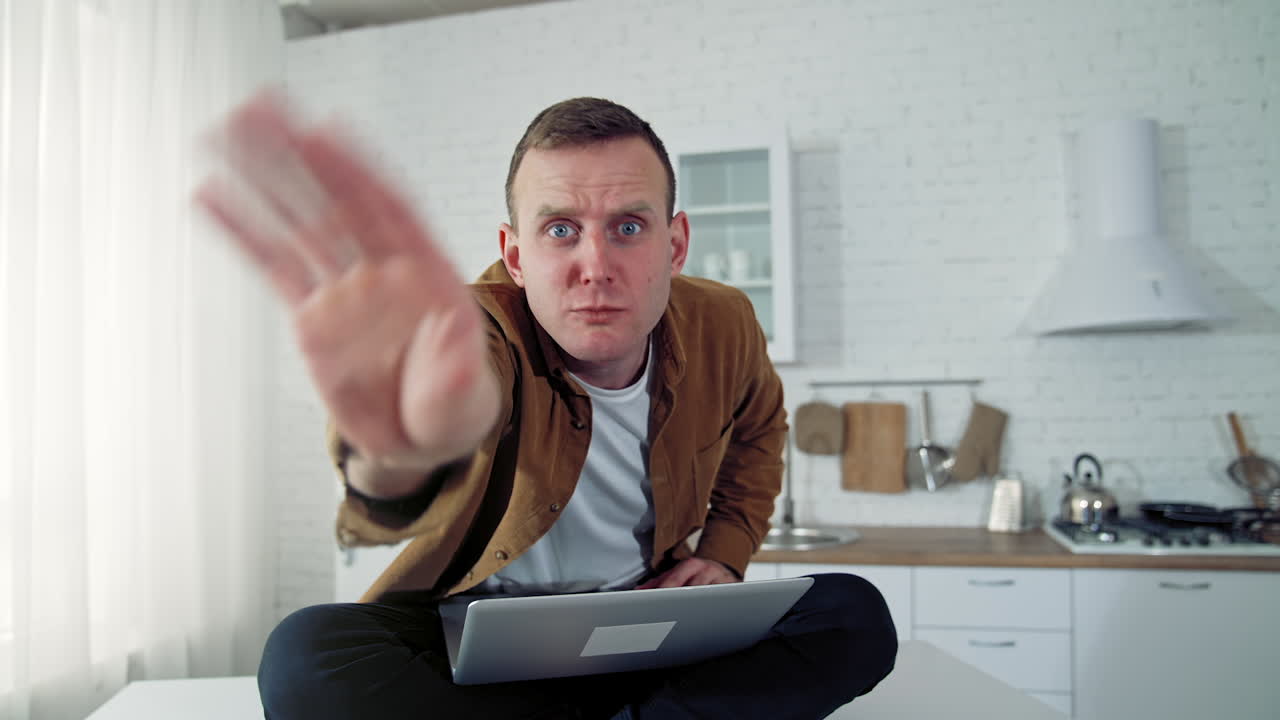 Funny comic businessman working at home. Mad face of a young freelancer looking on camera angrily while sitting on a kitchen table with a laptop.