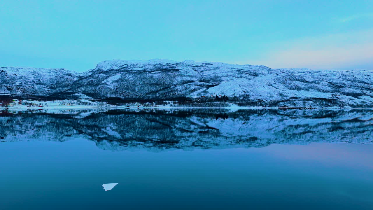 Betagende luftfoto af Lofoten-øhavet i Norge om vinteren, med snedækkede bjerge der perfekt reflekteres i fjordens rolige, blå vand ved skumringstid.