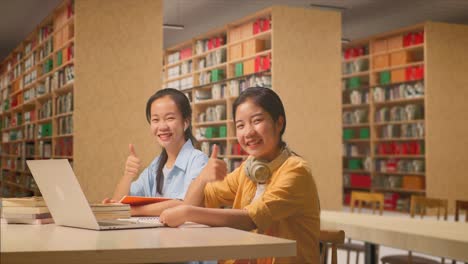 asian woman students with headphones smiling and showing thumbs up gesture to the camera while reading books on the table with laptop in the library