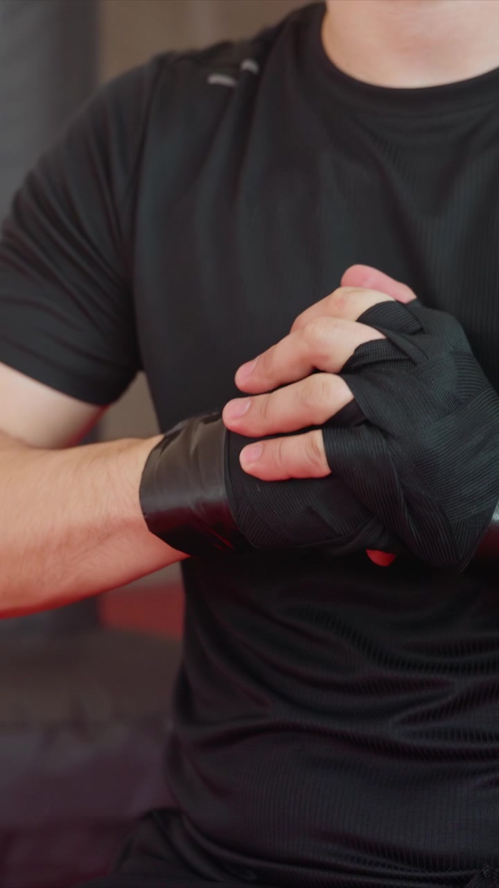 Primer plano de un hombre vendándose las manos con vendas negras. El proceso incluye asegurar la venda. Entorno interior con fondo oscuro, manteniendo el enfoque en la parte superior del cuerpo mostrando la mano del hombre.