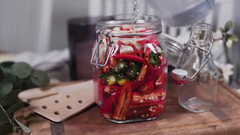 Vertiendo-Agua-Salada-En-Un-Frasco-De-Vidrio-Lleno-De-Ajo-Y-Chiles-Mixtos,-Habanero-Y-Carolina-Reaper,-Parados-En-Una-Tabla-De-Cortar-De-Madera-En-Una-Acogedora-Cocina-Con-Flores-Y-Utensilios-De-Cocina