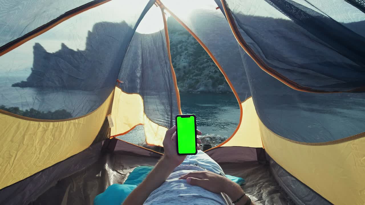텐트에 부착된 태양 전지판, 에 앉아있는 남자가 에서 드폰을 충전하고, 캠핑 텐트 안에 누워있는 젊은 남자가 녹색 화면의 스마트폰을 들고 해변에 서 있습니다.