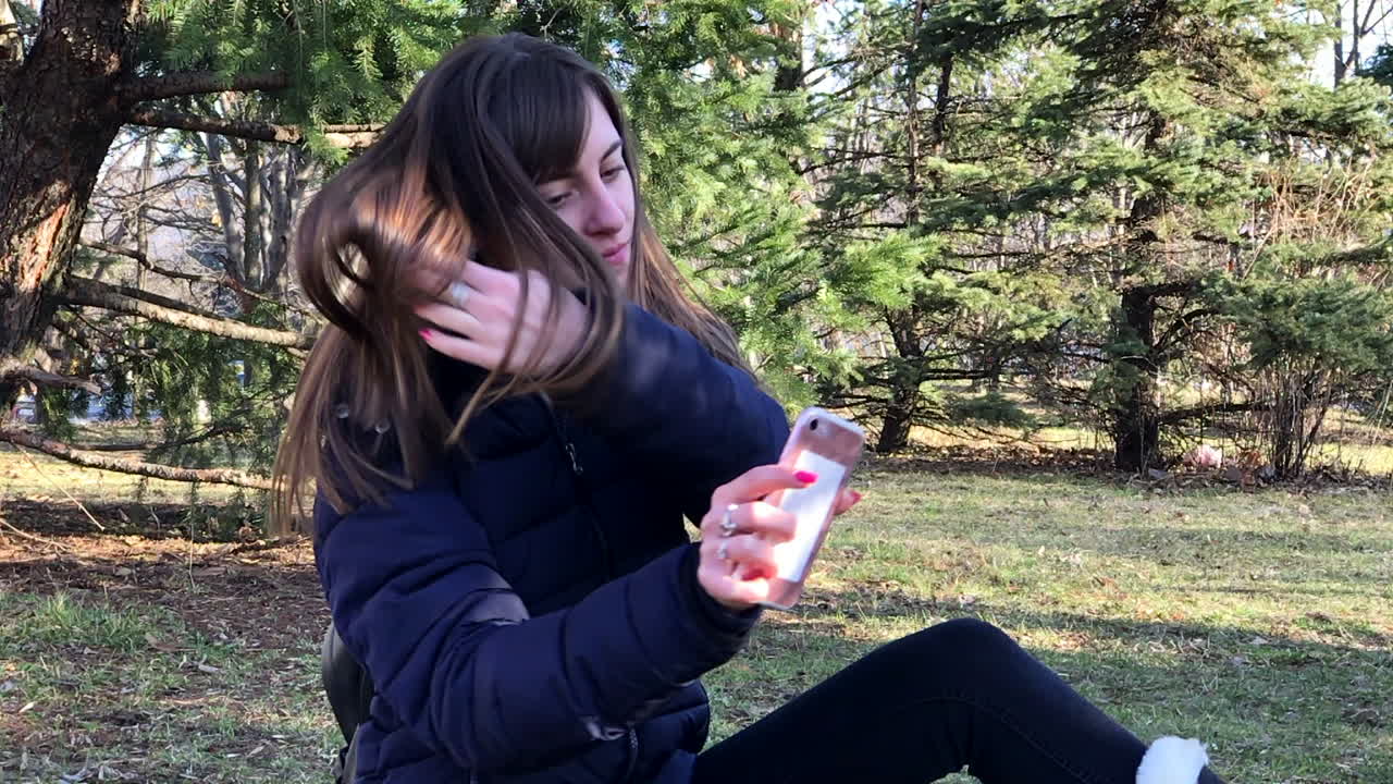 cámara lenta de una joven tomando selfies con un smartphone usando una cámara frontal sentada en el suelo en un parque urbano