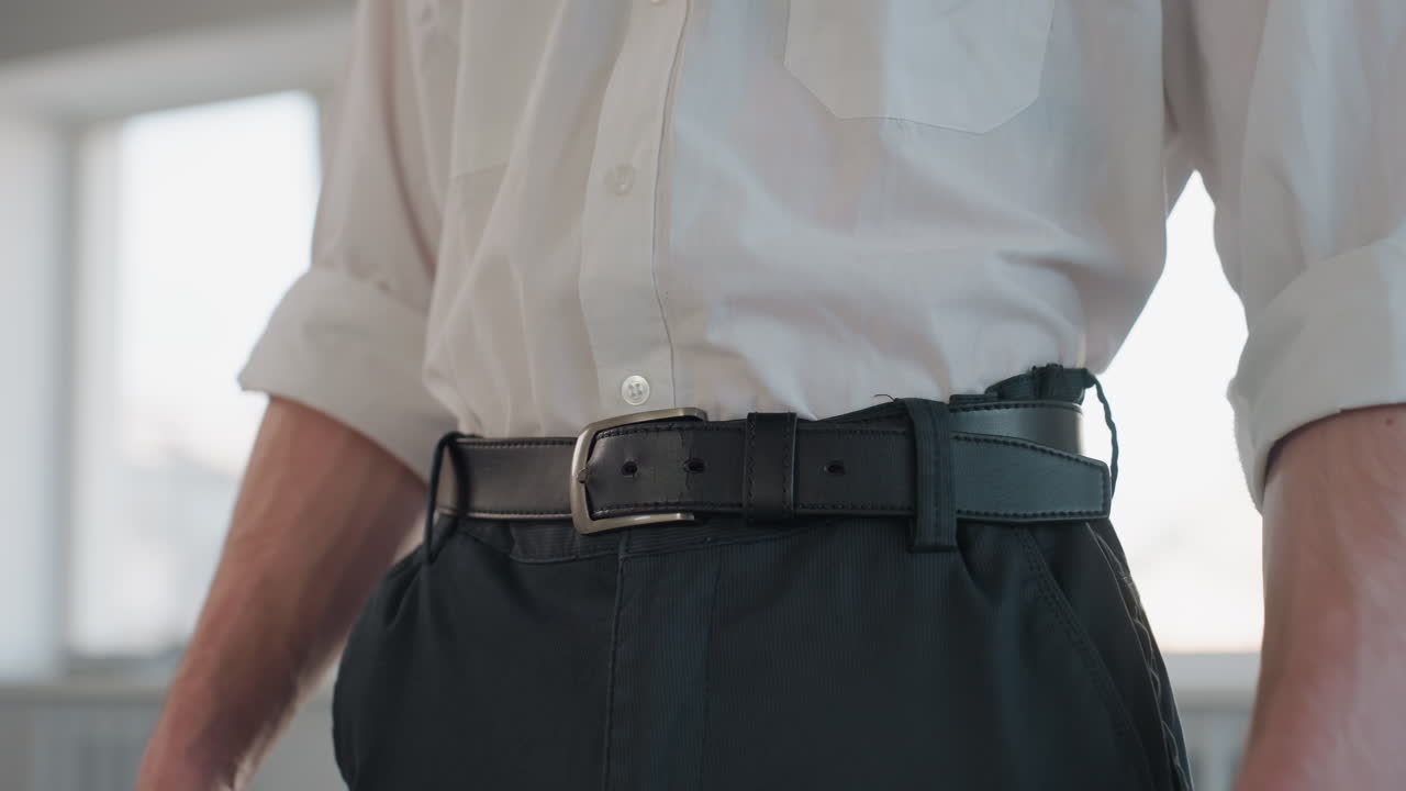 Portrait view showing young man hands unfastening black belt strap from buckle while wearing white shirt rolled sleeves and dark trousers against bright window backdrop conveying dressing transition