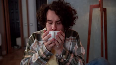 Anxious-brunette-guy-with-curly-hair-in-a-checkered-shirt-drinks-tea-from-a-white-cup-brings-the-cup-with-both-hands-to-his-lips-and-thinks-in-a-modern-apartment