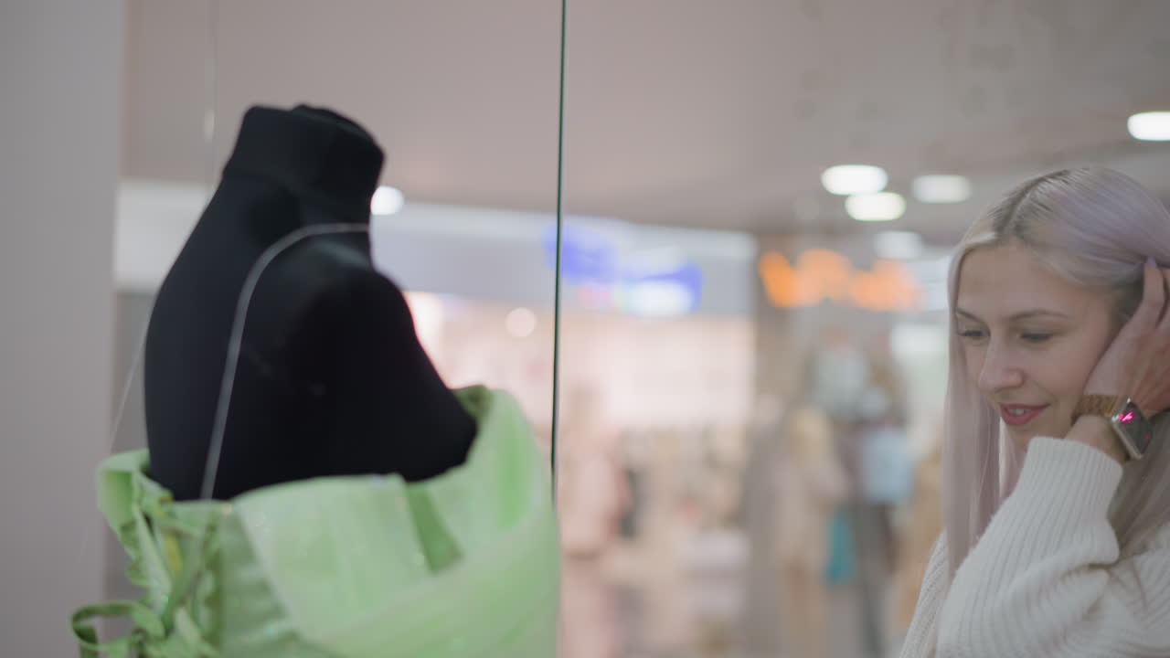 fair skinned lady bends to inspect green dinner gown behind glass with admiring smile, walking past boutique display, mannequins and shoppers visible in bright mall reflection lit by overhead lights
