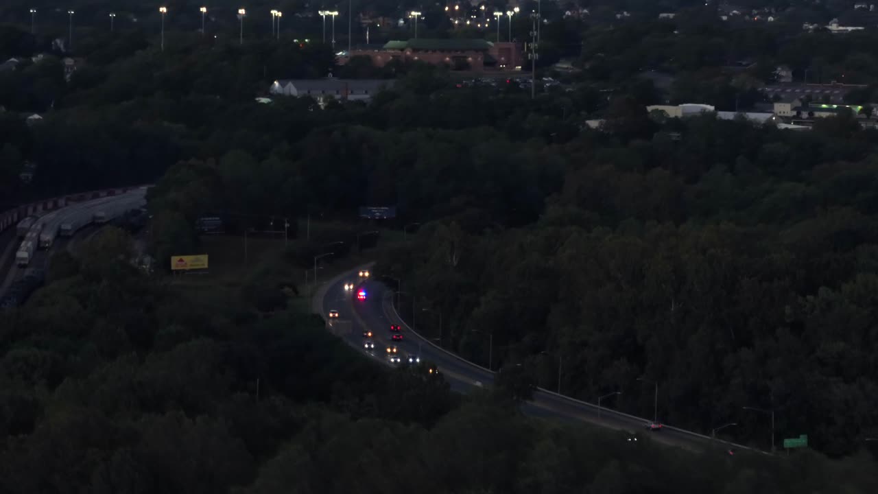 Police car with flashing lights on highway overtaking cars in suburbia. Aerial wide shot. Dusk scene in the evening. Forest trees in fall season. America, Virginia