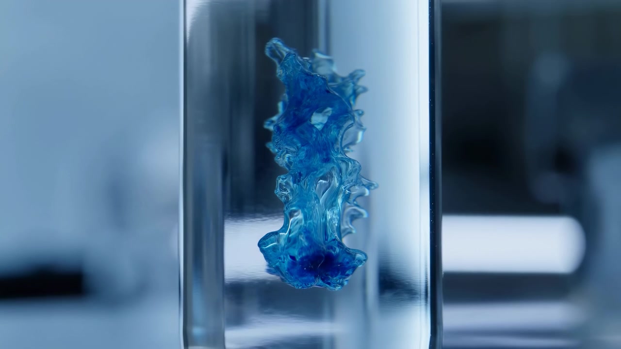 Un colore blu intenso si diffonde verso l'alto in delicate forme ramificate mentre un pigmento più denso rimane raccolto vicino alla base, incorniciato da vicino attraverso vetro trasparente su uno sfondo blu-grigio leggermente sfocato.
