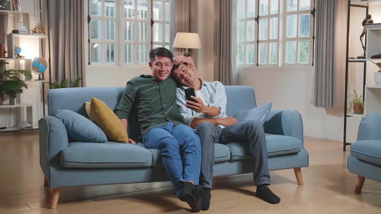 Asian Gentle Gay Couple Using Mobile Phone, While Sitting On A Couch In Cozy Stylish Apartment. Adult Boyfriends Online Shopping On Internet, Watching Funny Videos On Streaming Service