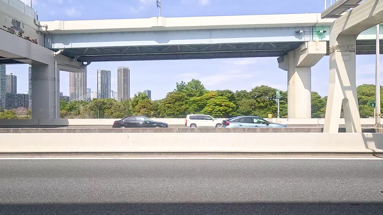 Vehicles move steadily on a sunlit Tokyo highway, framed by cityscape and greenery