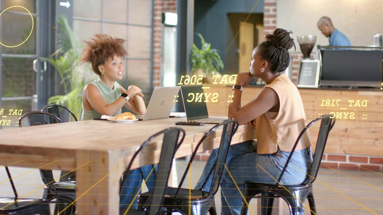 Two women gesturing and glancing at smartwatch, discussing business, yellow HUD overlaying laptops