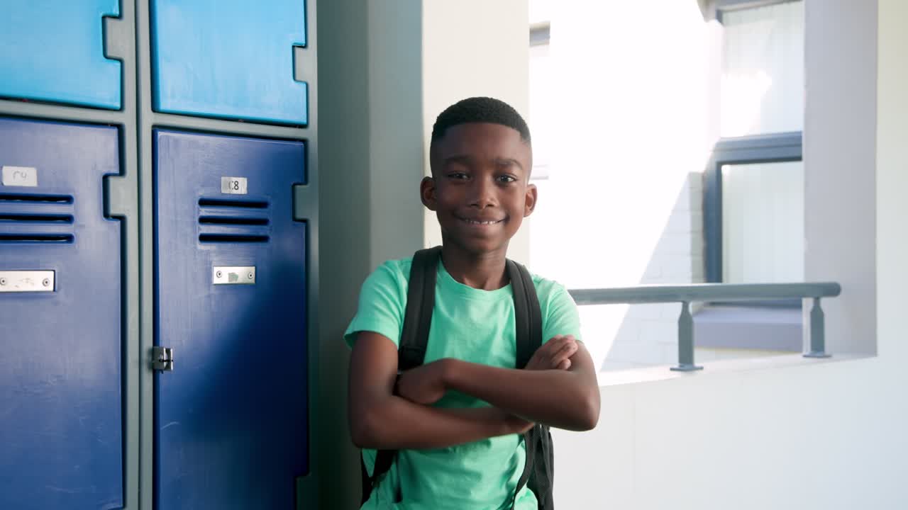 Entrando da destra, giovane ragazzo africano cammina verso gli armadietti blu a scuola, sorridendo, con le braccia incrociate e lo zaino.