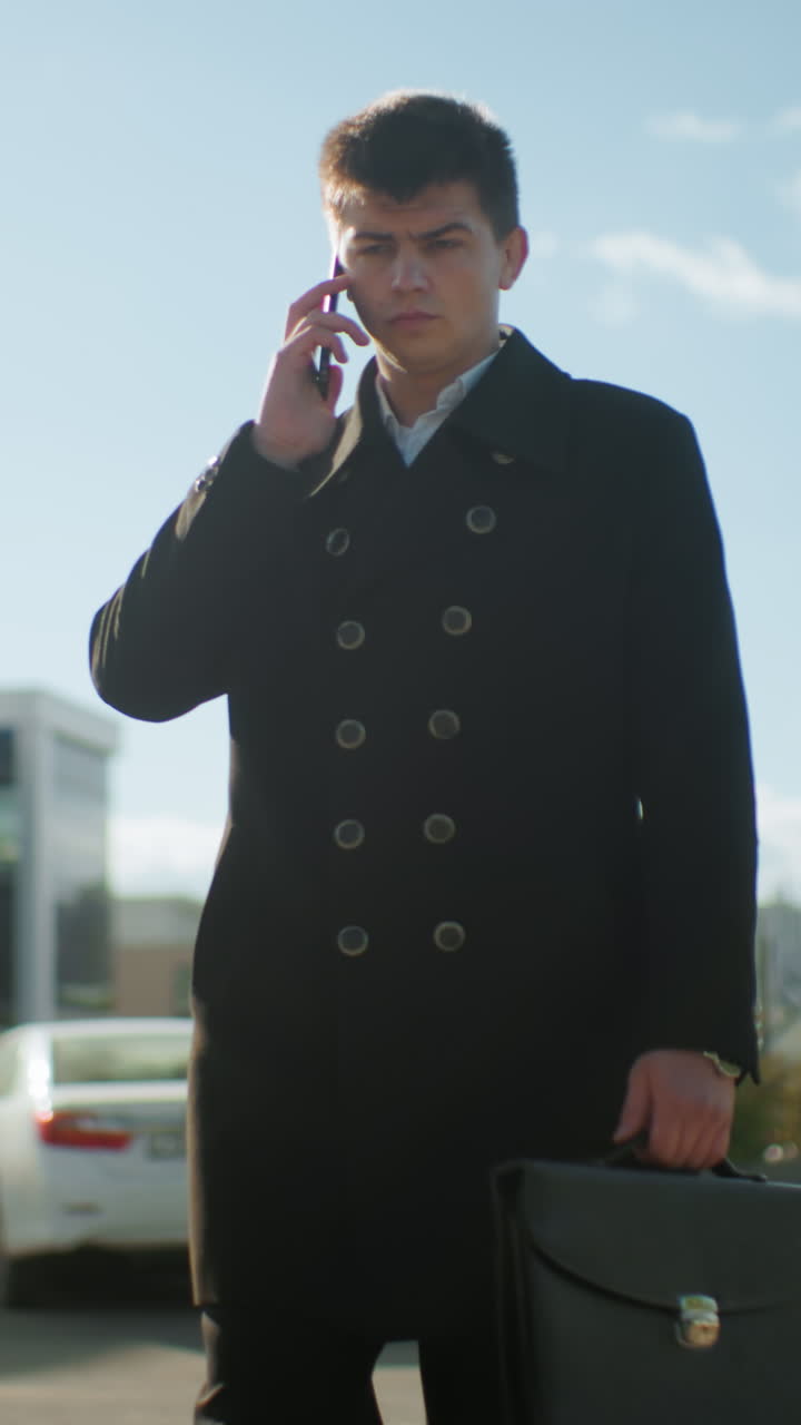 hombre de negocios en abrigo negro sostiene el teléfono bajo la mandíbula mientras continúa la llamada telefónica, caminando con confianza al aire libre bajo la luz del sol brillante en un entorno urbano cerca de coches estacionados y edificios modernos