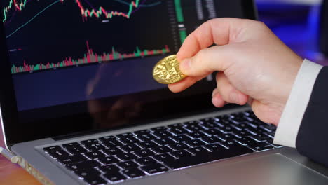 el hombre de negocios voltea bitcoin de oro en sus dedos mientras mira los gráficos de cambio y los precios en una computadora portátil