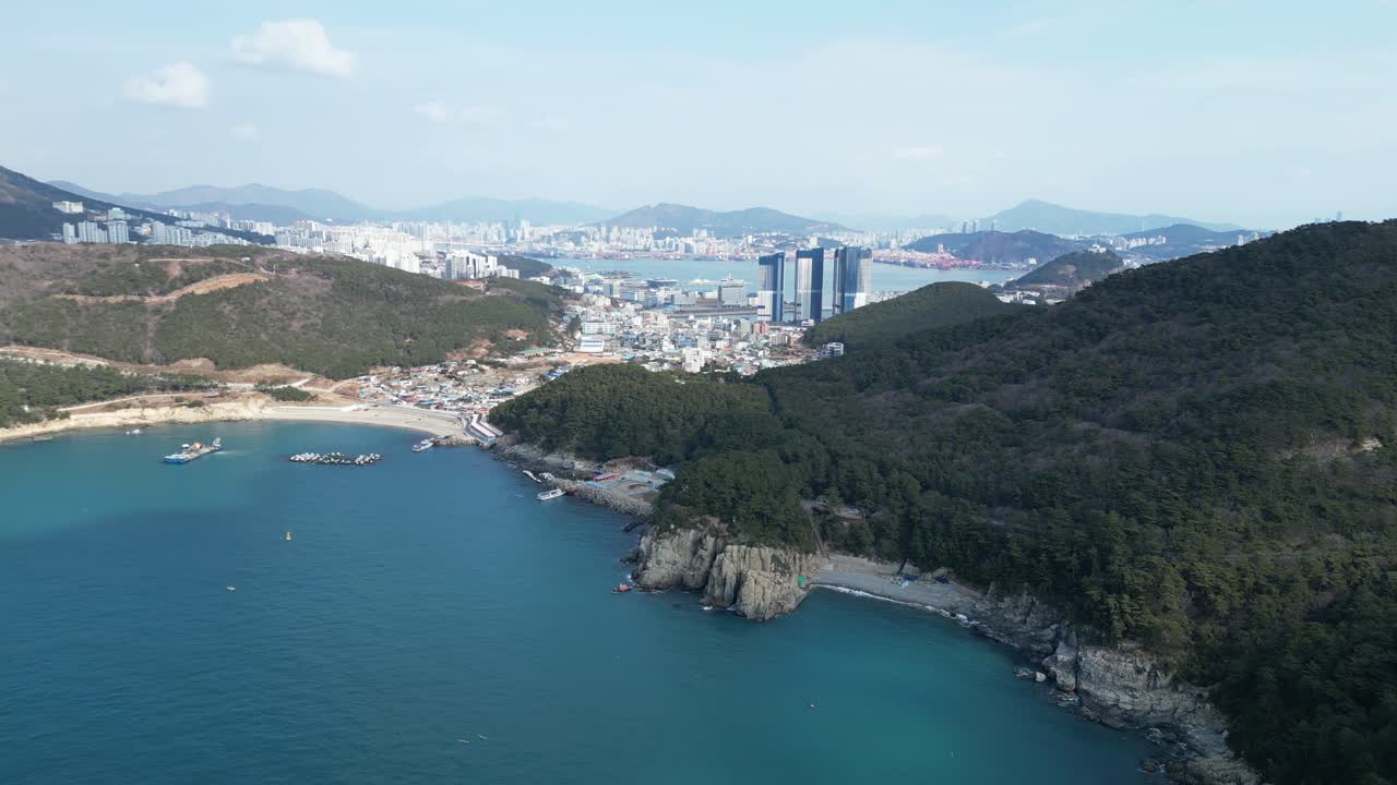 Drone aerial view in South Korea countryside flying over the dark blue sea of Busan next to a green mountain, ships and city in the background sunny day
