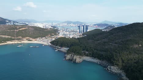 drone aerial view in south korea countryside flying over the dark blue sea of busan next to a green mountain, ships and city in the background sunny day
