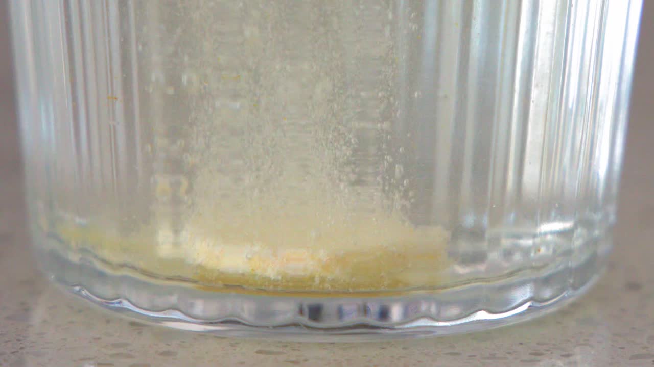 Primo piano di una compressa effervescente gialla che frigge e si dissolve in acqua dentro un bicchiere.