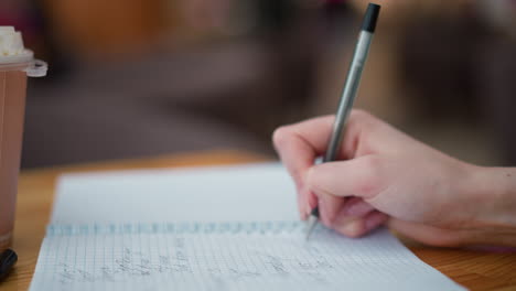 primer plano de escritura a mano con pluma negra en un cuaderno en una mesa de madera, cubierta de pluma al lado, y taza de café parcialmente visible, el fondo presenta una luz suave parpadeando