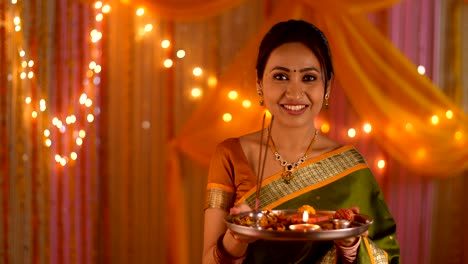 beautiful indian woman dressed in ethnic attire for festivities - holding a puja thali and smiling towards the camera