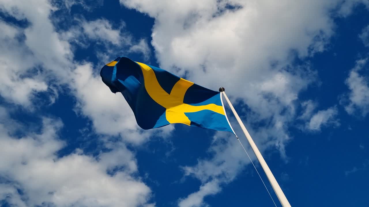 The Swedish flag sways gently in the breeze under a blue sky dotted with white clouds, filmed at Arlanda Airport near Stockholm, Sweden