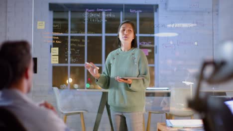 Asian-casual-businesswoman-using-tablet-and-glass-wall-presenting-to-colleagues-at-night-in-office