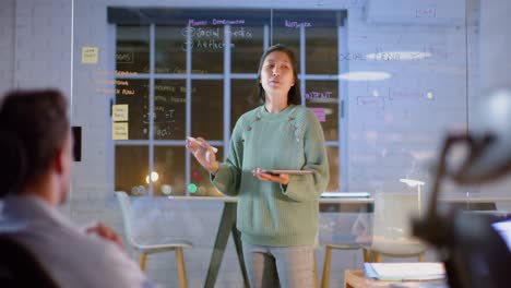 mujer de negocios asiática casual usando tableta y pared de vidrio presentando a sus colegas por la noche en la oficina