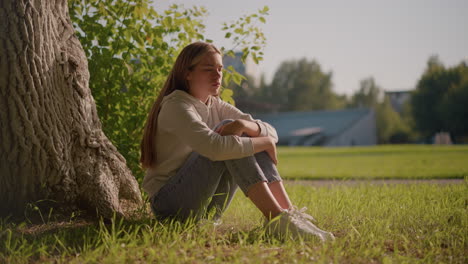 thoughtful woman sits on grassy ground, arms folded over knees, with sunlight softly illuminating her face, background features blurred poles and stadium