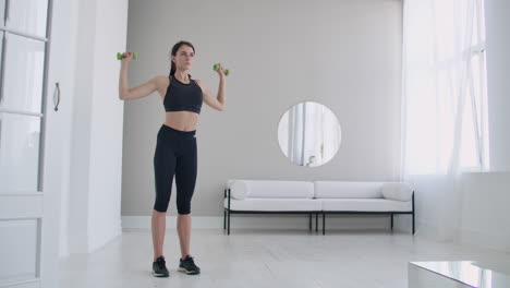 raise the dumbbells over your head performing exercises for the shoulders. training at home in the apartment. a brunette woman with long hair and a beautiful figure