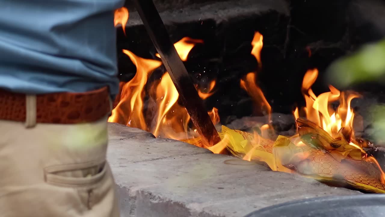 A person uses metal tools and paper to manage a fire in a concrete pit.